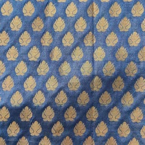 May include: A blue fabric featuring a repeating pattern of gold-coloured, leaf-shaped motifs arranged in a grid. The textile has a visible woven texture, and the metallic gold shapes are evenly spaced across the deep blue background surface.