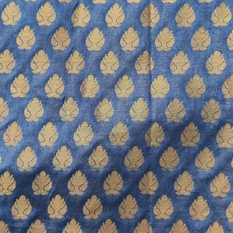 May include: A blue fabric featuring a repeating pattern of gold-coloured, leaf-shaped motifs arranged in a grid. The textile has a visible woven texture, and the metallic gold shapes are evenly spaced across the deep blue background surface.