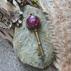 May include: A vintage-style key pendant with a round purple resin charm. The charm has a white swirl design and is set in a gold-toned metal frame.