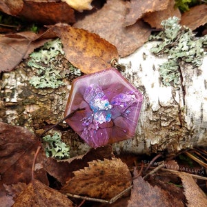 以下が含まれることがあります： 紫、茶色、白の抽象的なデザインが施された六角形のレジンペンダント。ペンダントは、葉と樹皮の自然な背景の上に置かれています。