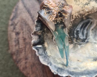 May include: A clear resin pendant with a gold compass design and a teal blue tail. The pendant is hanging from a brown chain and is resting on a wooden surface.