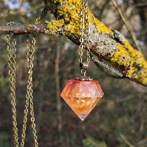 Puede incluir: Un collar con colgante en forma de diamante con una cadena de bronce cuelga de una rama de árbol. El colgante es de color naranja y rojo translúcido, con una parte superior decorativa de color bronce. El fondo está borroso.