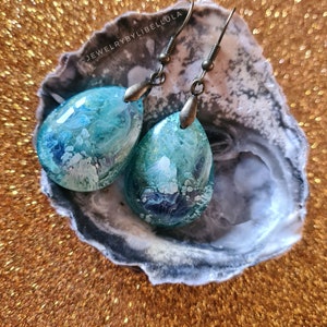 May include: A pair of teardrop-shaped earrings with a blue and white abstract design. The earrings are made of resin and have a bronze-colored hook.