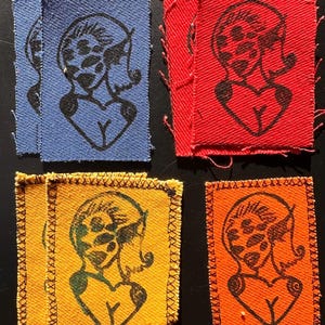 May include: A collection of fabric patches in blue, red, yellow, and orange. Each patch features a black line drawing of a woman's face and torso. The patches have a stitched border.