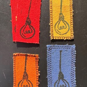 May include: Four rectangular fabric patches in red, yellow, orange, and blue. Each patch features a black illustration of a lightbulb with a hanging cord. The edges of the patches are stitched with a contrasting thread, adding a decorative border.