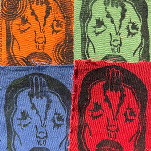 May include: Four fabric squares, each featuring a black linocut print of a stylized face with a hand above the head. The squares are in orange, green, blue, and red. The image has a handmade, artistic aesthetic.