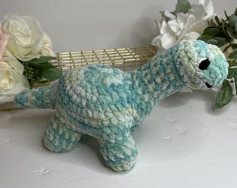 May include: A handmade crochet dinosaur in shades of blue, green, and white. The plush toy has a long neck, four legs, and a small tail. The dinosaur is sitting on a white surface, with flowers in the background.