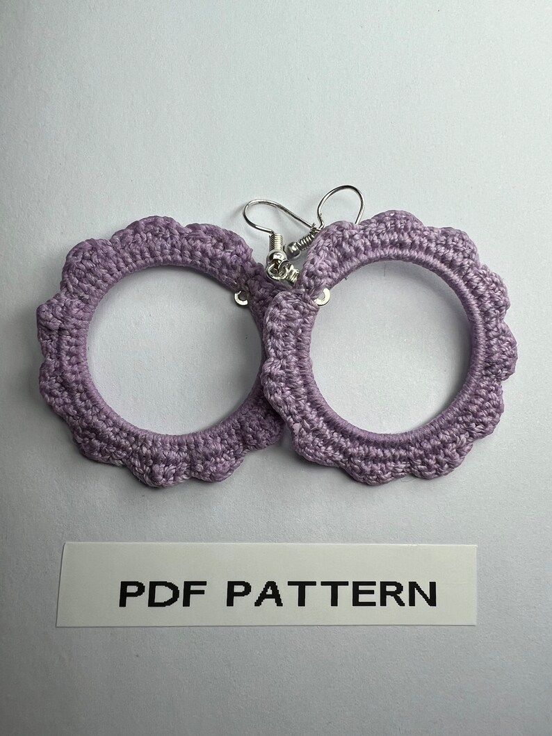 K&ouml;nnte beinhalten: Ein Paar geh&auml;kelte, lavendelfarbene Creolen mit silbernen Haken. PDF PATTERN steht auf einem wei&szlig;en Etikett unter den Ohrringen.