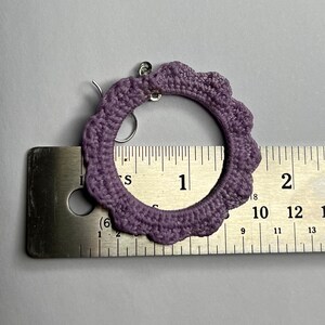 K&ouml;nnte beinhalten: Lilafarbene geh&auml;kelte Creolen mit einem silbernen Haken. Die Ohrringe werden an einem Lineal gemessen und zeigen, dass sie einen Durchmesser von etwa 2,5 cm haben.