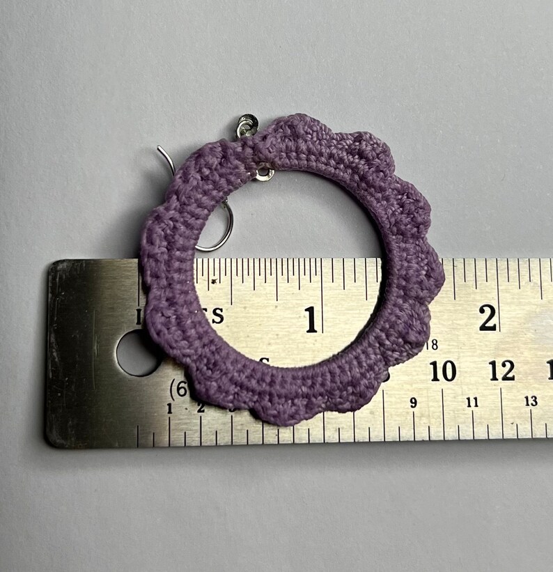 K&ouml;nnte beinhalten: Lilafarbene geh&auml;kelte Creolen mit einem silbernen Haken. Die Ohrringe werden an einem Lineal gemessen und zeigen, dass sie einen Durchmesser von etwa 2,5 cm haben.