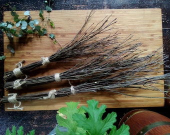 Può includere: Tre fasci di sottili ramoscelli marroni legati con spago, disposti su un tagliere di legno marrone chiaro. Sullo sfondo sono visibili foglie verdi e una botte di legno. I ramoscelli sembrano naturali.