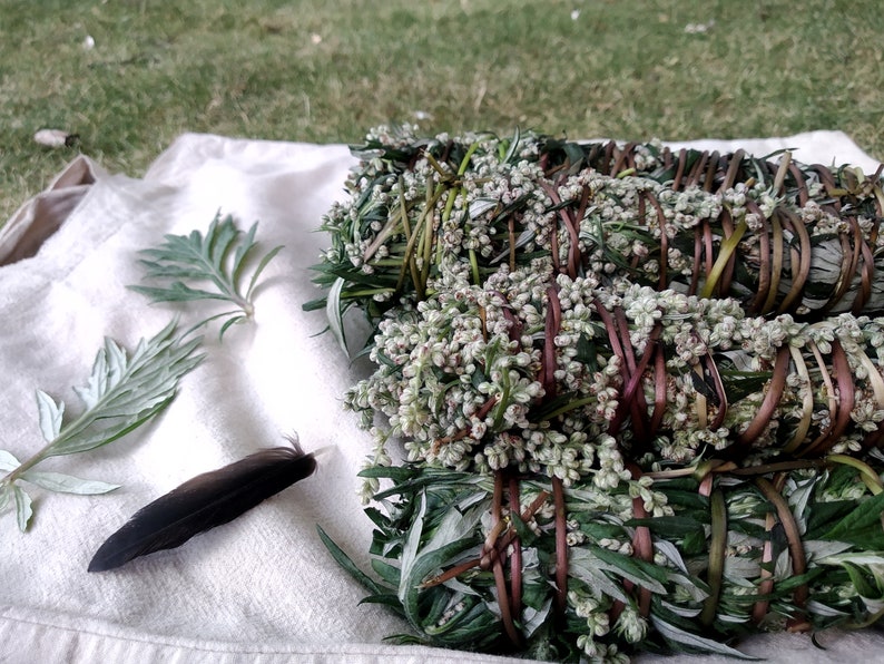 Pu&ograve; includere: Quattro mazzi di bastoncini di salvia secca legati con spago, disposti su un panno bianco con una singola piuma. La salvia &egrave; verde e grigia con piccoli fiori bianchi.