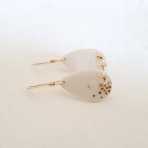 Peut inclure: Une paire de boucles d'oreilles en forme de larme blanche avec des accents de paillettes dorées. Les boucles d'oreilles sont en céramique et ont un fil d'or délicat.