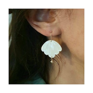 Peut inclure: Boucles d'oreilles en forme de coquille blanche avec un pendentif en or. Les boucles d'oreilles sont accrochées à l'oreille d'une personne.