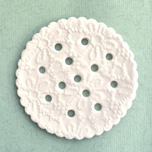 Puede incluir: Un posavasos blanco y redondo con borde festoneado y un patrón de encaje floral. El posavasos tiene 12 agujeros espaciados uniformemente. El fondo es un papel texturizado verde azulado claro.