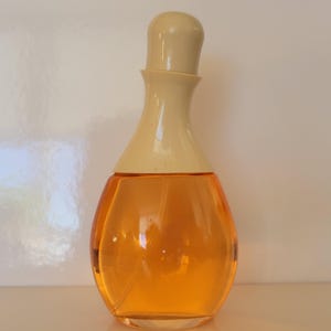 May include: A clear glass perfume bottle with an orange liquid inside. The bottle has a rounded shape with a cream-colored cap. The perfume bottle is sitting on a white surface.
