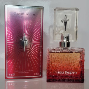 May include: A red and gold perfume bottle with a silver sword design on the cap. The bottle is labelled "Cesare Paciotti" and is 1.7 fl oz. The bottle is sitting on a white surface.