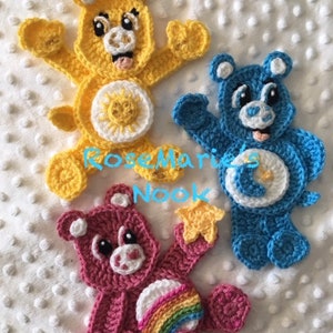 May include: Three crocheted Care Bear characters. The yellow bear has a sun symbol on its belly, the blue bear has a moon and star symbol, and the pink bear has a rainbow symbol.