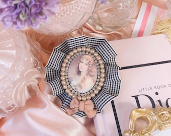 Black and white gingham checkered frill brooch with a printed classical-style female motif. Accented with a pearl teardrop and pink ribbon.