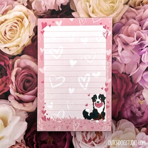May include: Pink notepad with lined paper and a white border of hearts. A cartoon Bernese Mountain Dog is holding a red heart in its mouth. The text "DUTCHDOGSTUDIO.COM" is at the bottom of the notepad.