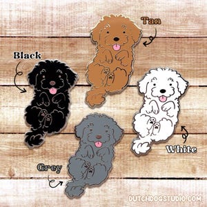 May include: Five enamel pins of a cartoon dog in different colours: black, grey, tan, white, and a light grey. Each pin has a pink tongue sticking out and the dog is sitting on its hind legs with its paws in the air. The pins are on a wooden background with the colour names written next to each pin.