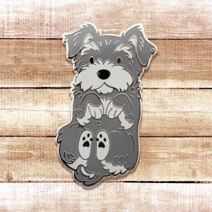 May include: A gray enamel pin of a miniature schnauzer dog sitting with its paws crossed. The dog has a white chest and a black nose.