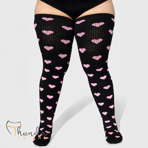 May include: Black thigh-high socks with a repeating pattern of pink hearts. The socks are ribbed and extend from the feet to mid-thigh. The background is a plain, light gray.