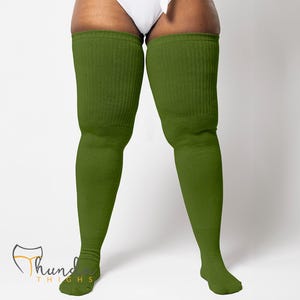 May include: Olive green thigh-high socks. The socks are made of a ribbed knit material and extend from the feet to the upper thighs. The socks are a solid color and appear to be comfortable and warm. The product is branded with the text "Thund Thighs".