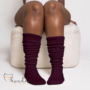 May include: A pair of burgundy slouch socks. The socks are ribbed and gathered at the ankles, with the text "Thunda Thighs" in a stylized font at the bottom left. The socks are worn by a person sitting on a white, textured ottoman.