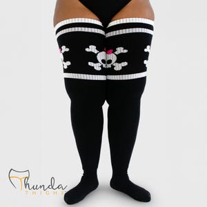 Puede incluir: Medias altas negras con rayas blancas en la parte superior. Las medias presentan un diseño de calavera y huesos cruzados blancos con un lazo rosa. La marca "Thunda Thighs" es visible en la esquina inferior izquierda.