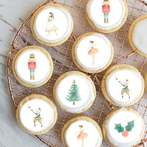May include: Round iced sugar cookies with watercolour illustrations of festive characters, including a ballerina, Christmas tree, and nutcracker. The cookies are arranged on a copper wire cooling rack. The cookies are a festive treat.