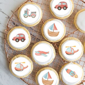 May include: Round iced biscuits decorated with watercolour illustrations of vehicles, including a car, scooter, aeroplane, sailboat, helicopter, and rocket. The biscuits are on a copper wire cooling rack.