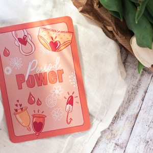 Könnte beinhalten: Eine rechteckige Karte mit pfirsichfarbenem Hintergrund und rotem Rand zeigt Illustrationen von Menstruationsprodukten und den Worten "Period Power". Die Karte enthält Bilder von einer Binde, Unterwäsche, Menstruationstassen und einem Tampon. Die Karte liegt auf einem weißen Tuch mit Tulpen.