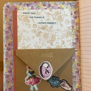 K&ouml;nnte beinhalten: Offenes Notizbuch mit floral gemusterten Seiten und einem braunen Umschlag. Der Umschlag tr&auml;gt die Aufschrift "UR MAGIC" und Aufkleber eines Schlosses, einer Frau in einem Kleid und Blumen. Eine Karte mit dem Text "KNOW THAT THE TIMING IS ALWAYS PERFECT" ist ebenfalls sichtbar.