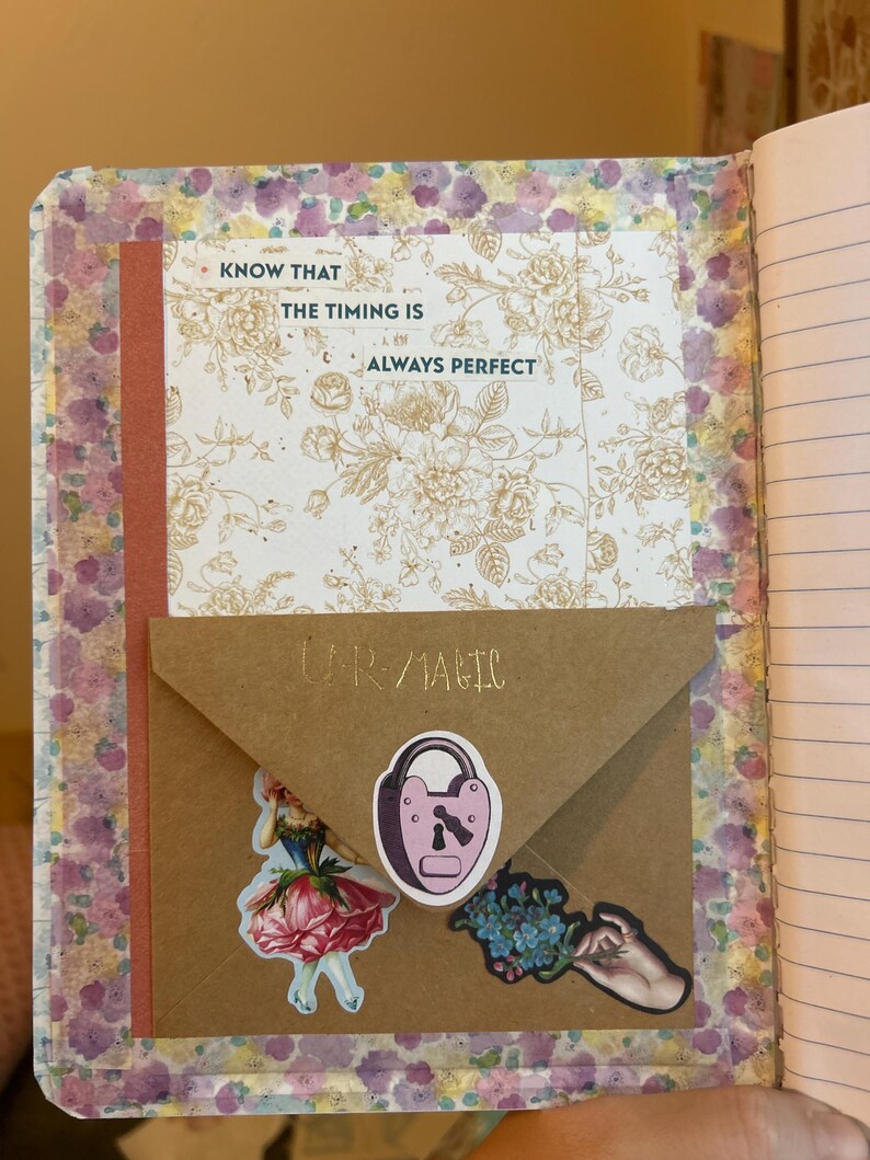 K&ouml;nnte beinhalten: Offenes Notizbuch mit floral gemusterten Seiten und einem braunen Umschlag. Der Umschlag tr&auml;gt die Aufschrift "UR MAGIC" und Aufkleber eines Schlosses, einer Frau in einem Kleid und Blumen. Eine Karte mit dem Text "KNOW THAT THE TIMING IS ALWAYS PERFECT" ist ebenfalls sichtbar.