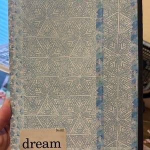 K&ouml;nnte beinhalten: Ein Notizbuch mit hellblauem und wei&szlig;em geometrischem Muster auf dem Einband. Der Einband tr&auml;gt das Wort "dream" auf einem rechteckigen Etikett. Der Buchr&uuml;cken ist schwarz, und die Kanten sind leicht abgenutzt.
