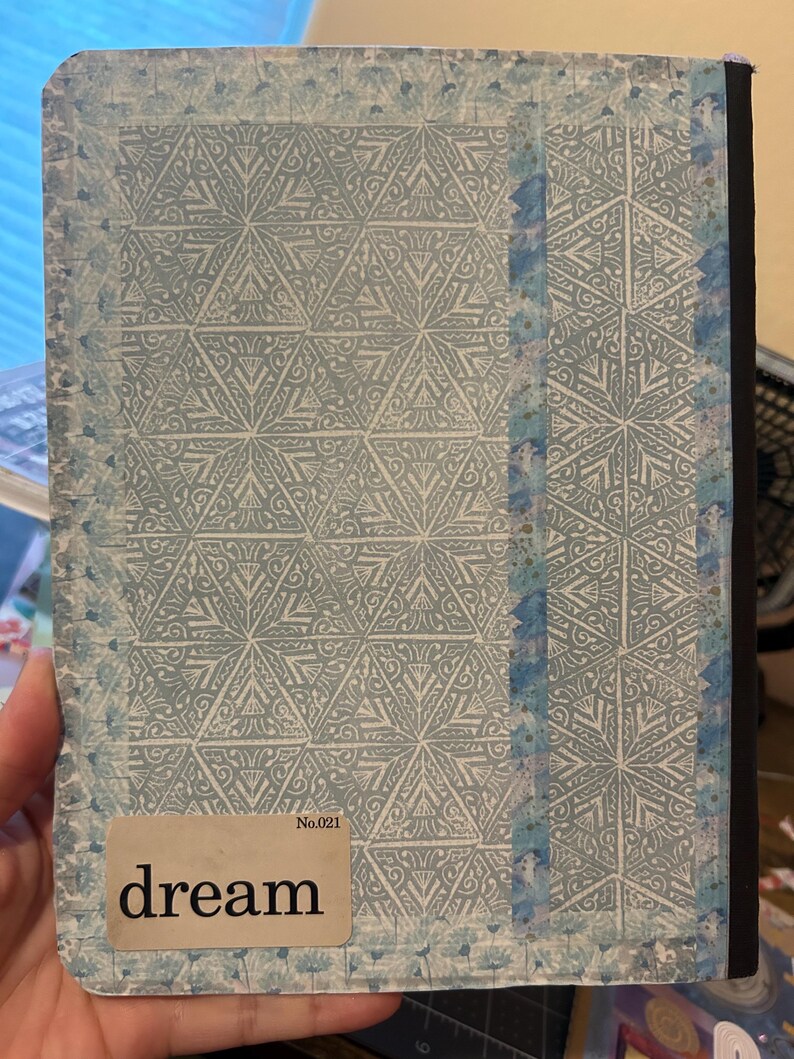K&ouml;nnte beinhalten: Ein Notizbuch mit hellblauem und wei&szlig;em geometrischem Muster auf dem Einband. Der Einband tr&auml;gt das Wort "dream" auf einem rechteckigen Etikett. Der Buchr&uuml;cken ist schwarz, und die Kanten sind leicht abgenutzt.