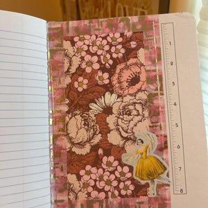 K&ouml;nnte beinhalten: Offenes Notizbuch mit linierten Seiten und einem dekorativen Blumendesign. Das Design zeigt einen braunen Hintergrund mit rosa und cremefarbenen Blumen, akzentuiert mit goldenem Folienband und einem Aufkleber einer Frau in einem gelben Kleid. Ein Lineal ist auf der rechten Seite sichtbar.