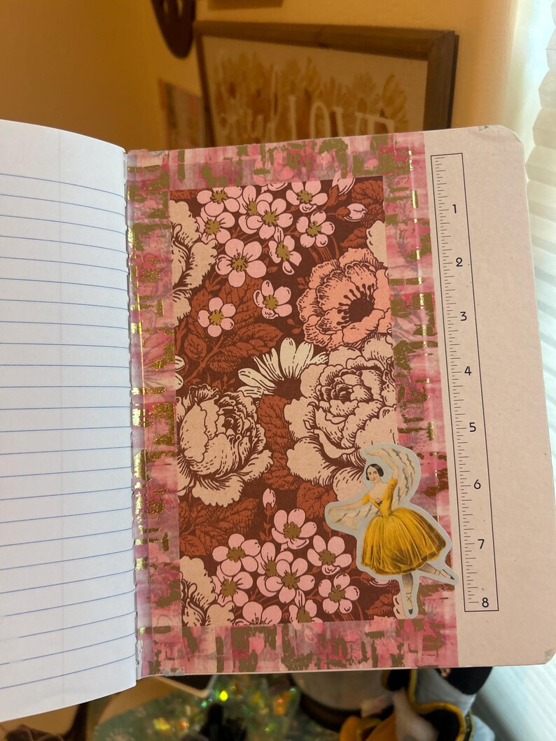 K&ouml;nnte beinhalten: Offenes Notizbuch mit linierten Seiten und einem dekorativen Blumendesign. Das Design zeigt einen braunen Hintergrund mit rosa und cremefarbenen Blumen, akzentuiert mit goldenem Folienband und einem Aufkleber einer Frau in einem gelben Kleid. Ein Lineal ist auf der rechten Seite sichtbar.