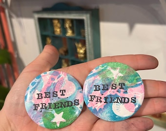 Könnte beinhalten: Zwei runde Buttons mit dem Text "BEST FRIENDS" in Schwarz. Die Buttons haben ein farbenfrohes Design mit rosa, blauen und grünen Farbtönen. Ein Button zeigt einen weißen Stern.