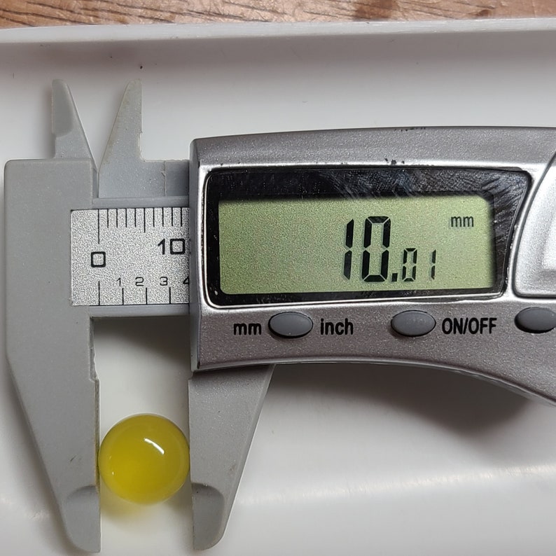 May include: A yellow, spherical object is measured with a digital caliper. The caliper displays a measurement of 10.01 mm. The object is positioned on a white surface.