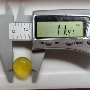 May include: A digital caliper measuring a translucent yellow sphere. The digital display shows a measurement of 11.97 mm. The sphere is held between the caliper jaws, indicating its diameter.