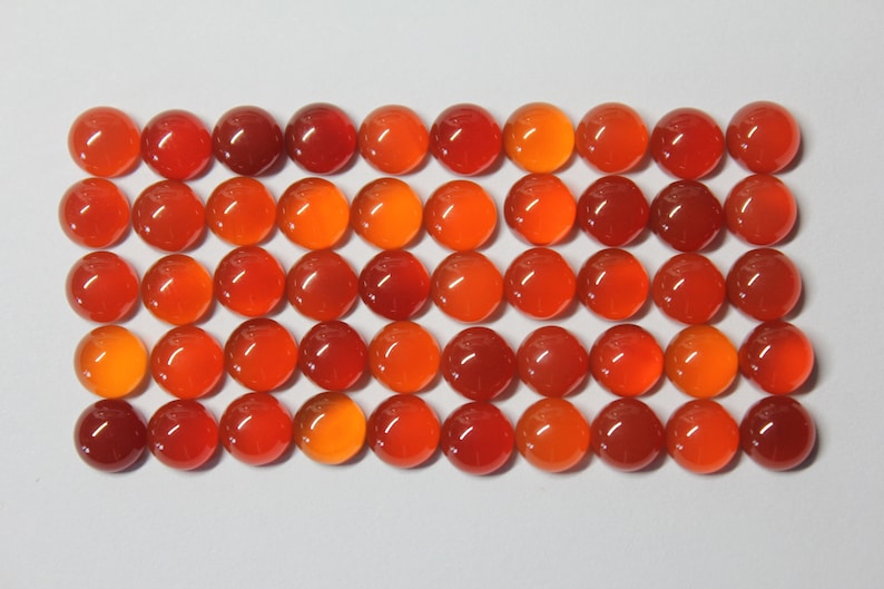 Puede incluir: Una colecci&oacute;n de 40 cabujones redondos de cornalina naranja. Los cabujones est&aacute;n dispuestos en una cuadr&iacute;cula sobre una superficie blanca.