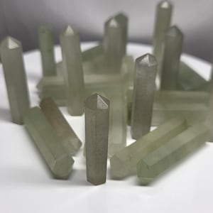 May include: A collection of pale green crystal points, each with a hexagonal base and a pointed top. The crystals are arranged on a white surface, with some standing upright and others lying down. The crystals have a translucent appearance.