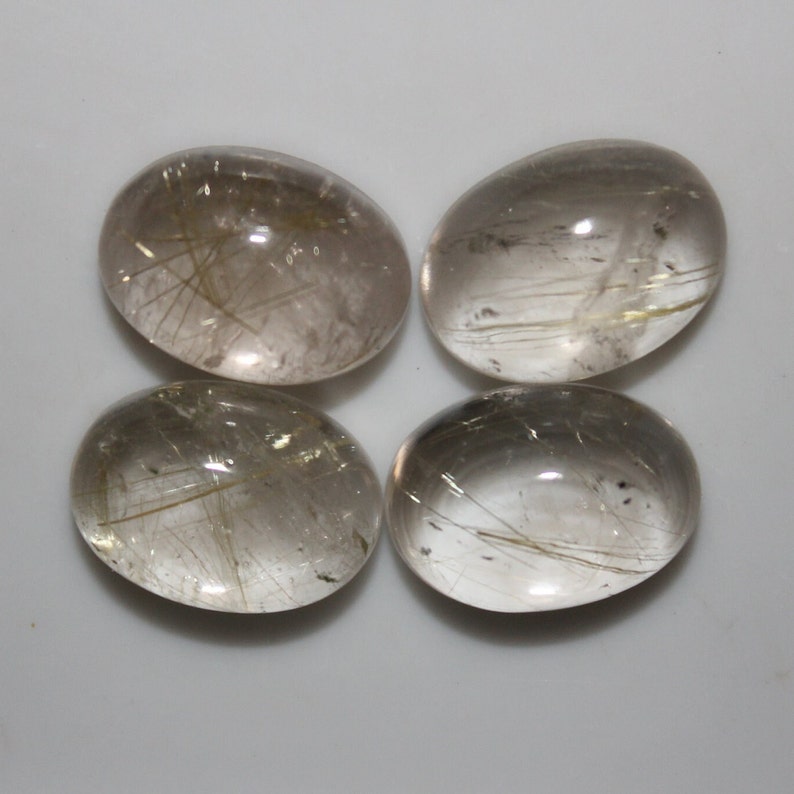May include: Four oval-shaped, translucent, clear quartz cabochons with gold rutile inclusions. The cabochons are smooth and polished.