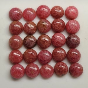 May include: Twenty-four round, polished gemstones in shades of pink and red are arranged in a grid pattern. The gemstones have a smooth, reflective surface and vary in color intensity, from light pink to deep red-brown.