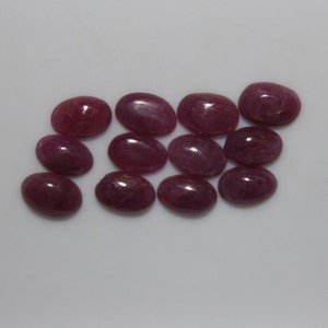 May include: A collection of 12 oval-shaped, pink ruby gemstones. The gemstones are smooth and polished, with a slight shimmer.