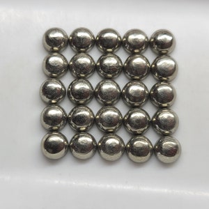 May include: A collection of 25 round, silver-colored pyrite stones arranged in a grid pattern. The stones are smooth and have a metallic sheen.