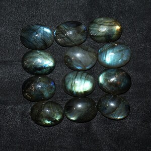 May include: Twelve oval-shaped labradorite gemstones with iridescent blue and green hues. The stones are arranged in three rows of four.