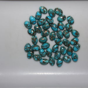 May include: A collection of oval-shaped turquoise gemstones with a copper-colored matrix. The stones are scattered across a white surface. These polished stones are suitable for jewelry making.
